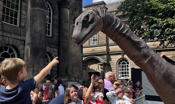 photo of a child reaching upwards from a crowd to touch the nose of a life-sized dinosaur roaming the streets